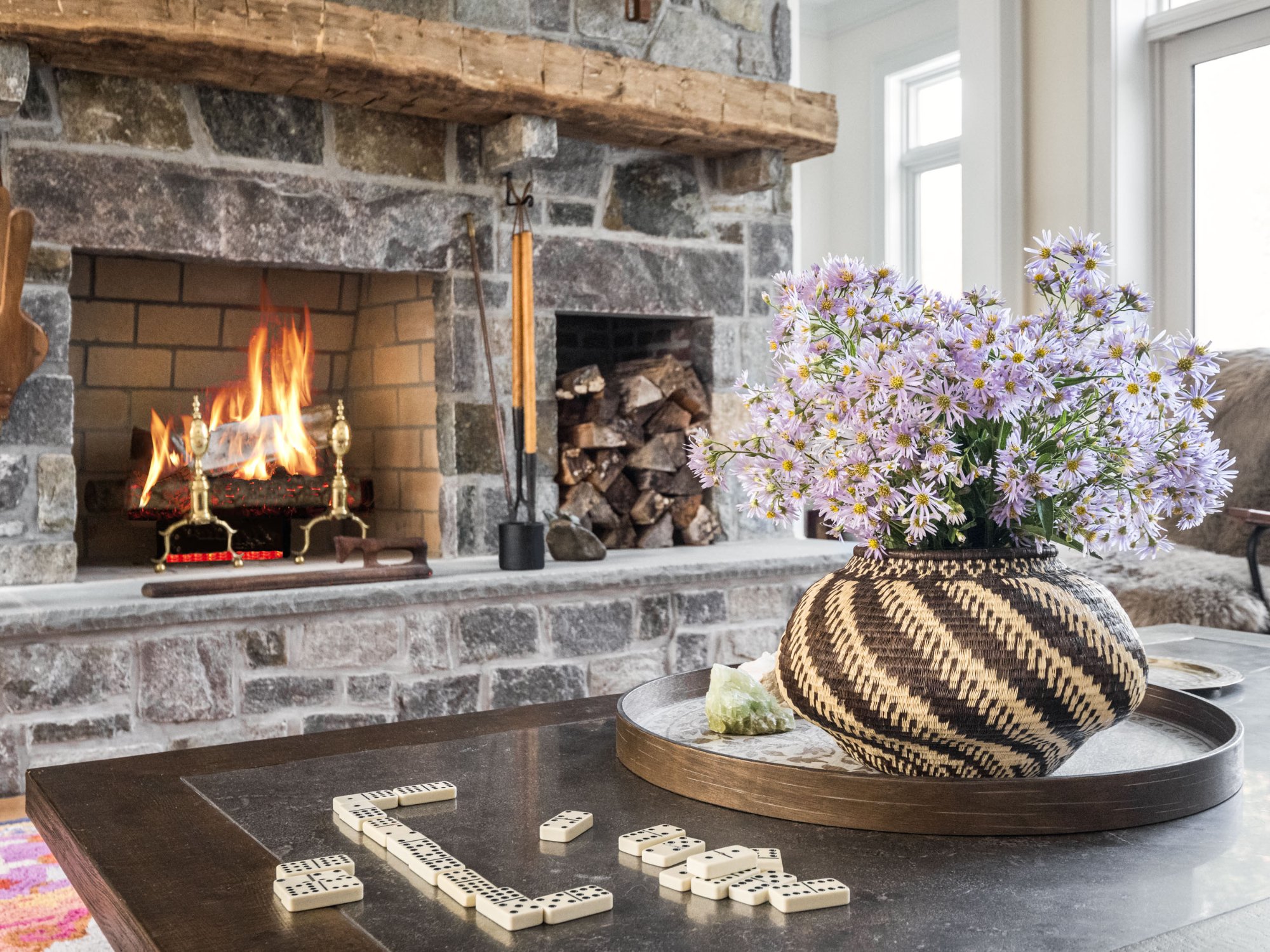 Coffee table detail with dominoes, woven vase with wildflowers, and stone fireplace with burning fire — Nordic Farmhouse